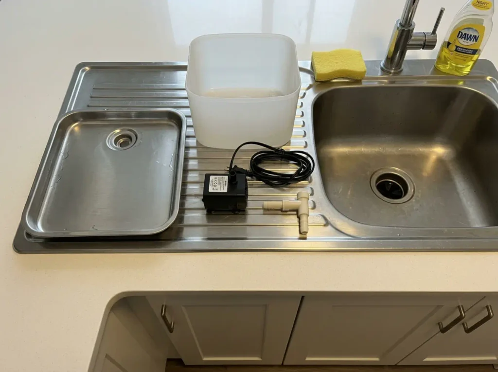 Disassembled cat fountain by the sink showing a steel tray beside a plastic reservoir and pump