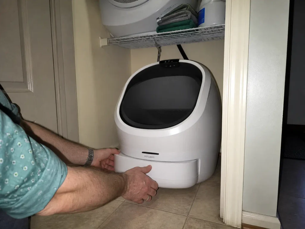 Hands sliding the waste drawer out of an open-top automatic litter box in a laundry alcove