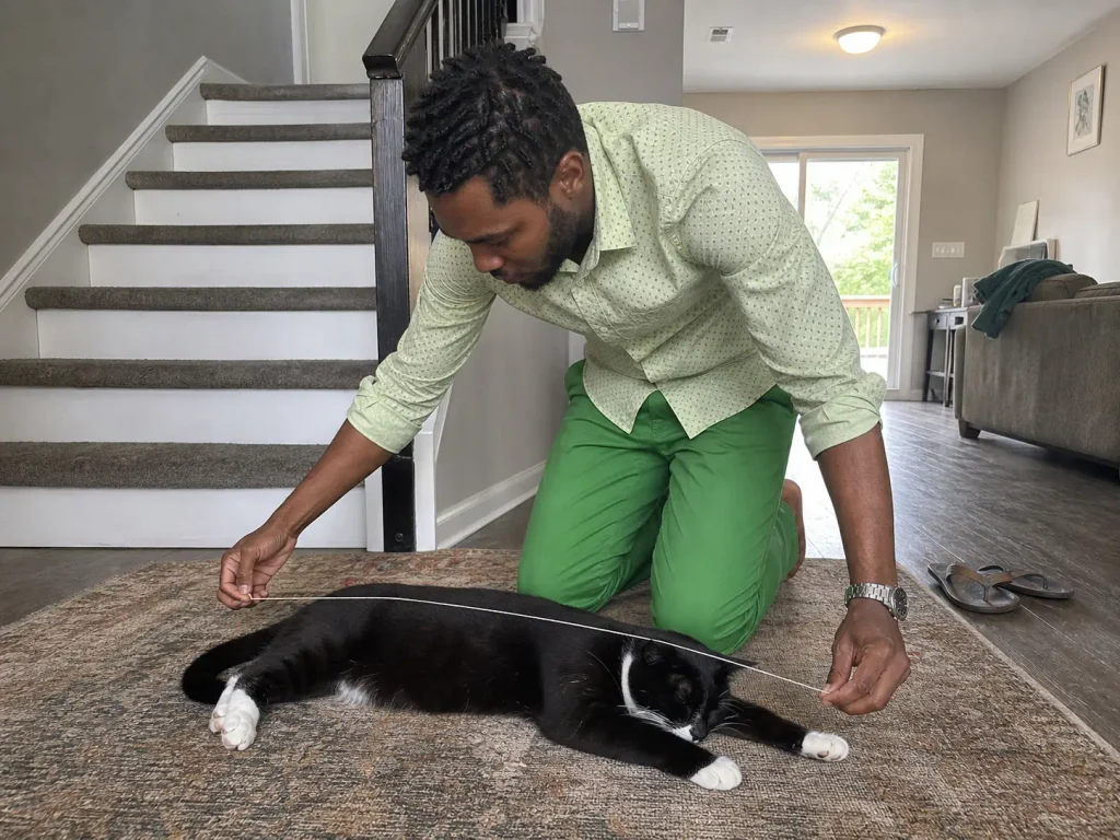 Man kneeling on a living room rug holding a length of string alongside a sleeping tuxedo cat