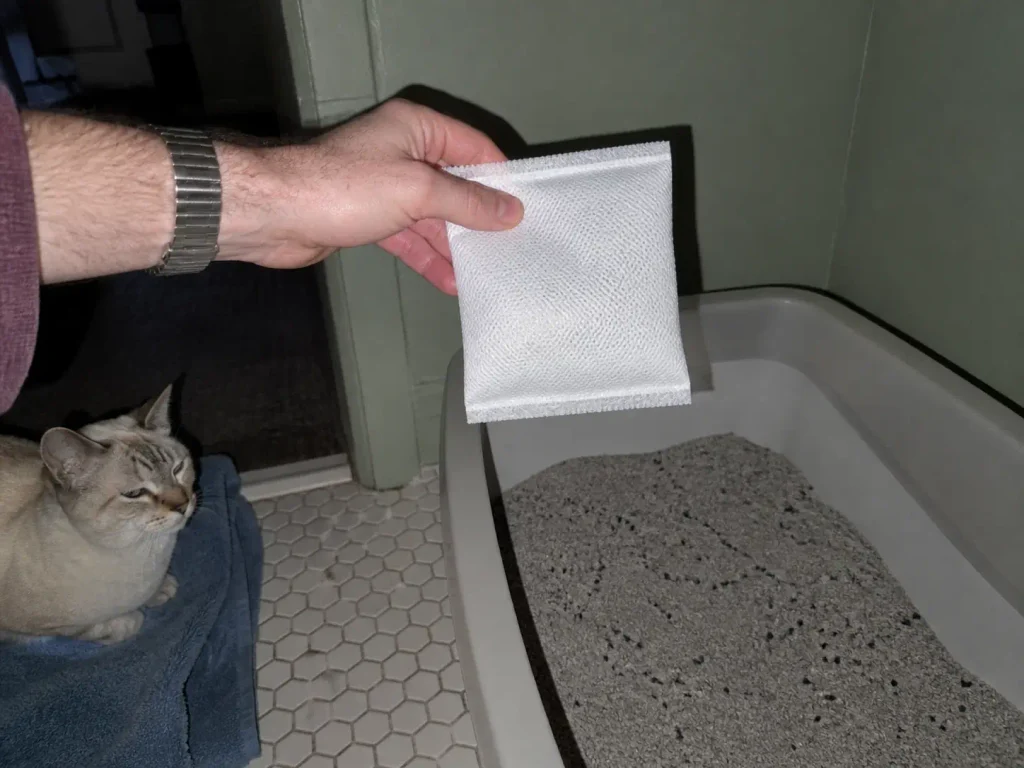 Activated charcoal pouches held in a hand above an open litter box on a bathroom floor