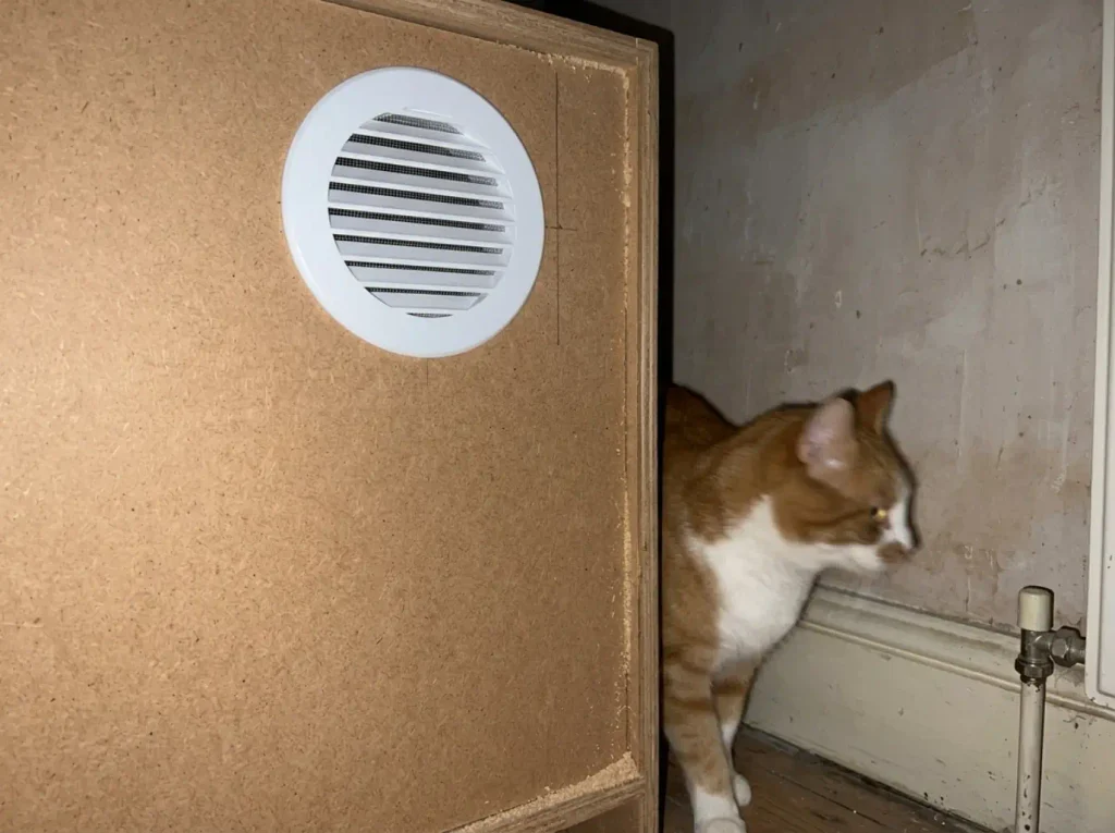 Round mesh vent cover fitted high on the back panel of a DIY litter box cabinet, seen from behind