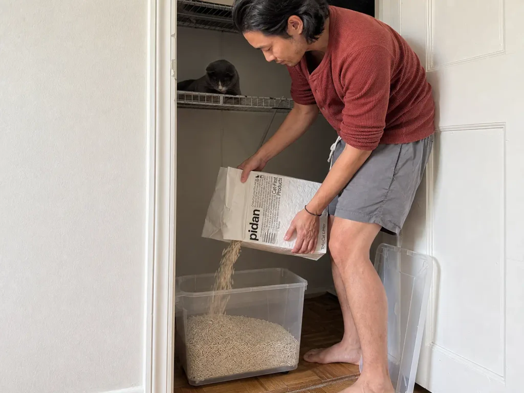 A bag of tofu cat litter being tipped into a sealed storage bin inside a hallway closet