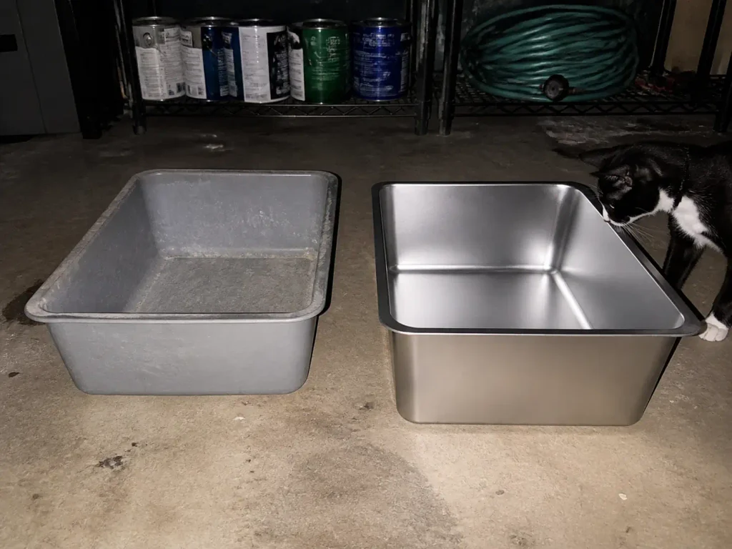 Old scratched plastic litter box next to a new stainless steel one on a garage floor