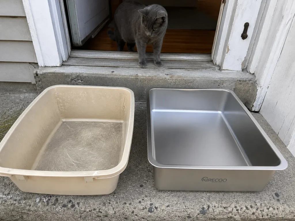 A new stainless steel litter box on a porch step beside an old scratched plastic one