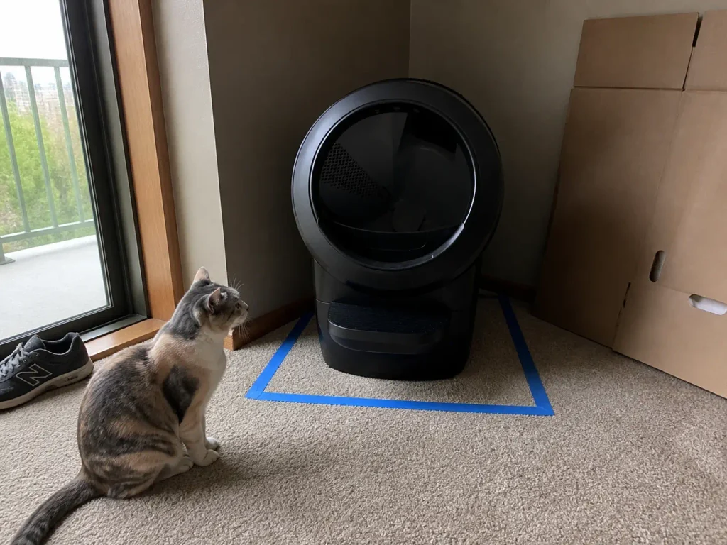 Automatic litter box in a living room corner with painter's tape marking its footprint on the floor