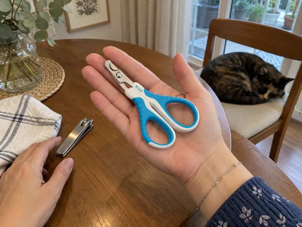 A hand holds a small closed scissor-style cat nail clipper above a dining table next to a human nail clipper